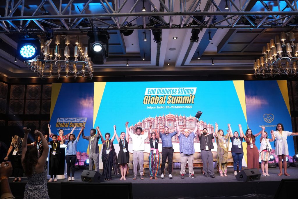 A wide shot of a brightly lit stage at the "End Diabetes Stigma Global Summit" in Jaipur, India. A diverse group of about fifteen people stand in a line across the stage, smiling and holding hands high in celebration. The large digital backdrop behind them is vibrant blue and yellow, featuring the event title and a prominent illustration of the Hawa Mahal palace. Above the stage, professional event lighting rigs and decorative chandeliers are visible, while a few audience members in the foreground cheer them on.