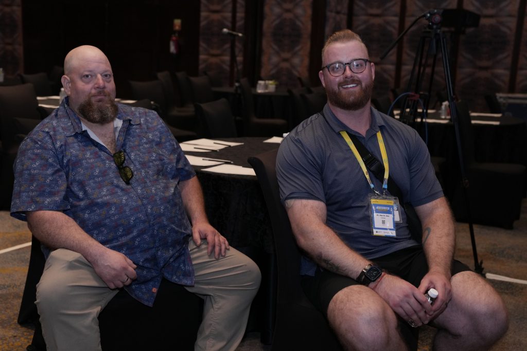 Two men sitting at a round conference table in a dimly lit banquet or seminar hall. The man on the left has a shaved head, a full brown beard, and is wearing a blue patterned button-down shirt with khaki pants. The man on the right is smiling, wearing glasses, a grey polo shirt, black shorts, and an event lanyard with a yellow strap around his neck. The background shows empty black chairs, additional tables, and a camera tripod setup.