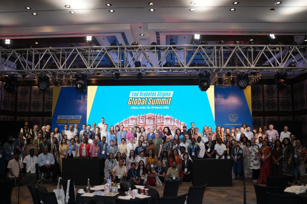 A wide, high-angle group photograph of a large, diverse crowd of attendees gathered in front of the main stage at the "End Diabetes Stigma Global Summit" in Jaipur, India. Dozens of people of various backgrounds and ages stand and kneel together, smiling for the camera. Behind them, the large digital screen displays the event title in white and yellow font against a bright blue background, featuring an illustration of the Hawa Mahal palace. The top of the frame shows an intricate metal lighting truss with stage lights hanging over the ballroom.