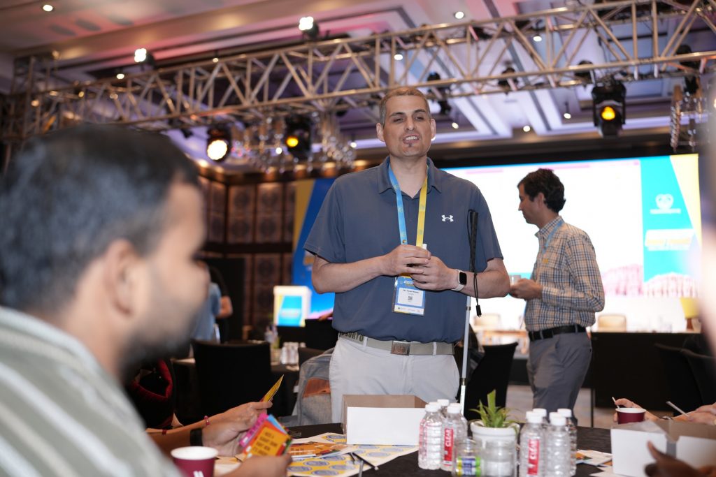 A medium shot focusing on a man with short grey hair speaking while standing at a conference table. He is wearing a grey Under Armour polo shirt, light-colored pants, a white smartwatch, and an event lanyard around his neck, and he is holding a white cane. In the blurred foreground, other attendees sit at the table, which holds water bottles and small cards. In the background, another man in a plaid shirt stands near the main stage area, which features lighting trusses and a large digital screen.
