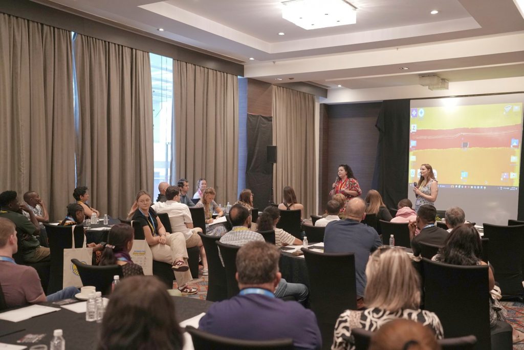 ALT for Image: Wide view of a room at the pre-summit film screening discussion, with facilitators speaking to attendees gathered around tables throughout the room.