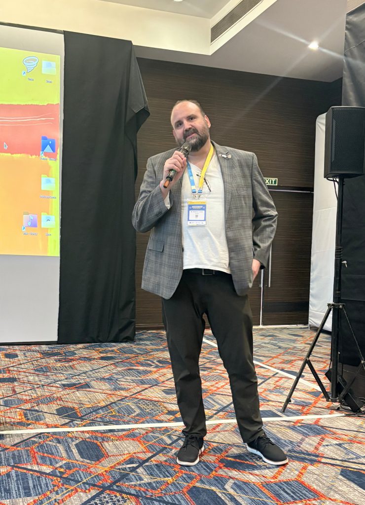 Matt Larson stands wearing a grey blazer at the front of a conference room, holding a microphone and responding to audience questions after the screening of Low Priority. 