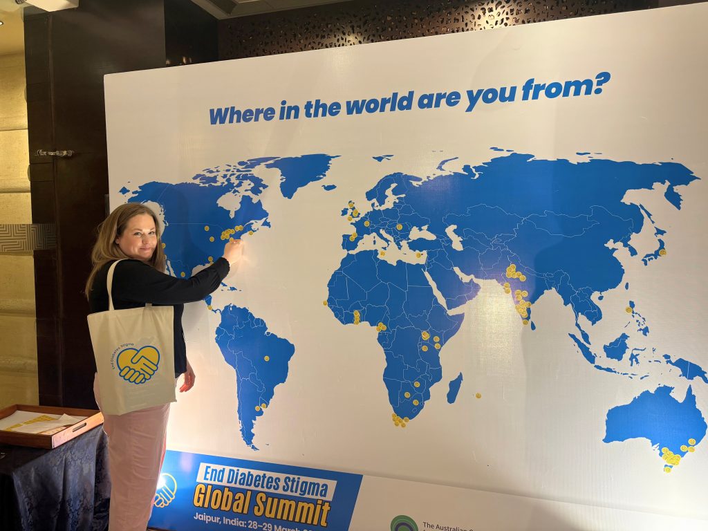 Brit Hancock standing in front of the map of the world. She points to Toronto, Canada on the map while smiling at the camera. 