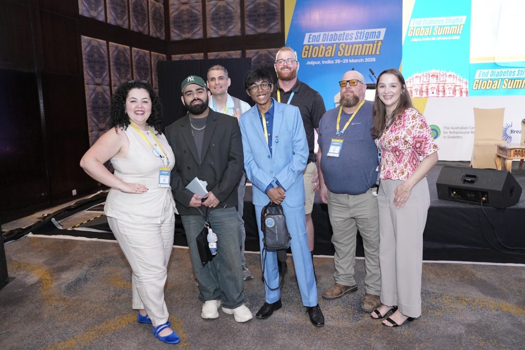 A group of seven diverse individuals smiling and posing together at an indoor event. Behind them is a large stage backdrop featuring a graphic of the Hawa Mahal and text that reads "End Diabetes Stigma Global Summit, Jaipur, India: 28-29 March 2026." The attendees are dressed in a mix of smart-casual and formal attire, wearing event lanyards.
