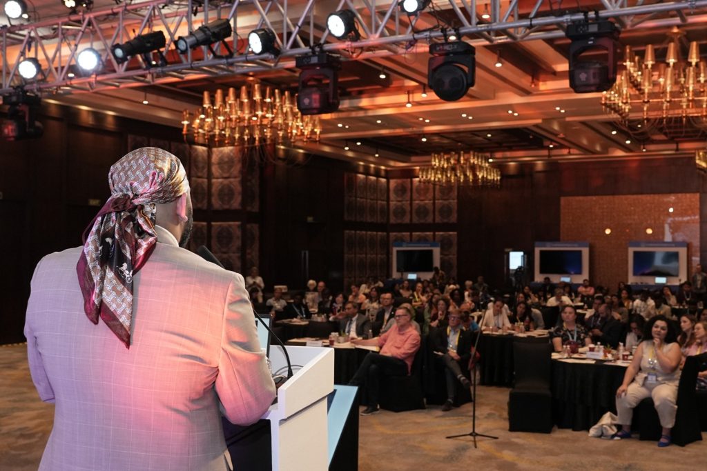 An over-the-shoulder shot from behind Anmol standing at a white podium, facing a large, seated audience in a grand ballroom. The speaker is wearing a light-colored plaid blazer and a patterned silk headscarf tied at the back. The audience sits at round tables, listening attentively. Overhead, heavy metal stage trusses hold professional event lighting and spotlights beneath a warm, ornately lit ceiling with chandeliers.