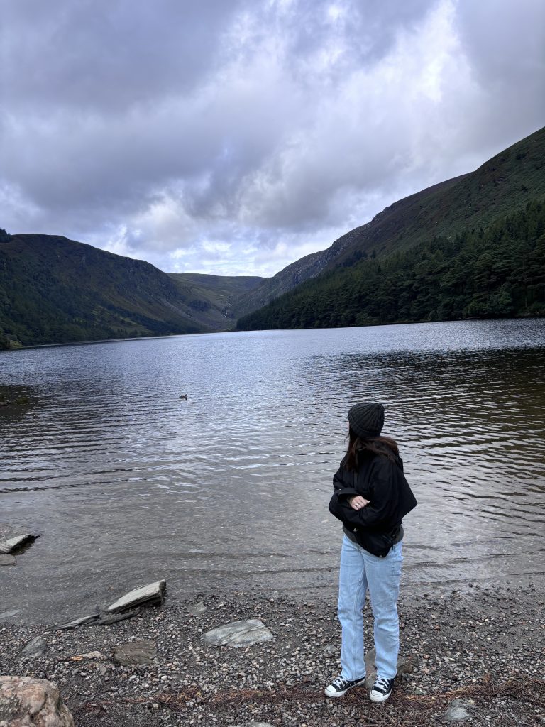 A scenic landscape photograph of a Macenzie standing on a rocky shoreline, viewed from the side as she looks out over a large, calm lake. She is dressed warmly in light blue jeans, a black jacket, and a dark beanie. The lake is surrounded by steep, green, tree-covered hills under a dramatic, overcast gray sky.
