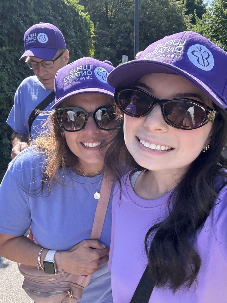 A close-up outdoor selfie of Macenzie and her mom and dad at the Lupus Ontario walk. All three are wearing matching purple baseball caps featuring the "Lupus Ontario" logo and the slogan "Life Without Lupus." The two women are also wearing sunglasses and lavender t-shirts, capturing a bright, sunny day at a community walk or event.