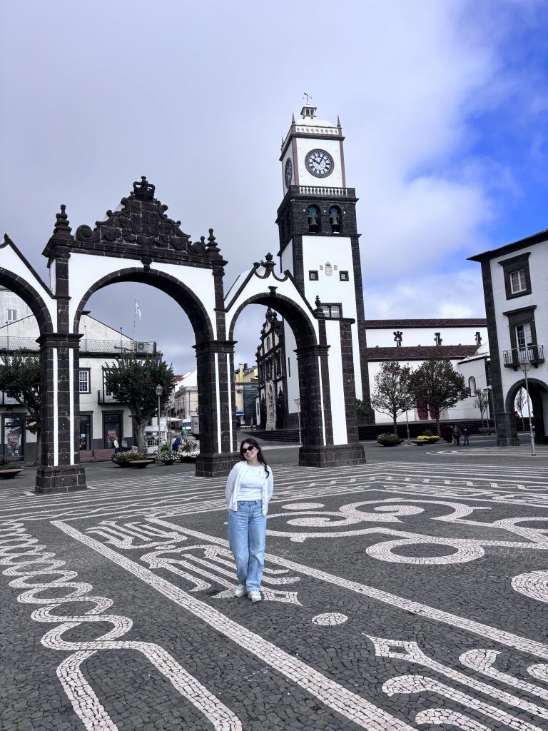 A wide shot of a Macenzie standing in a Praça Gonçalo Velho Cabral square, paved with intricate black and white mosaic patterns. She is smiling and wearing light-wash jeans and a white cardigan. Behind her stands a large, ornate three-arched stone gateway, with a white clock tower and church building set against a partly cloudy blue sky.