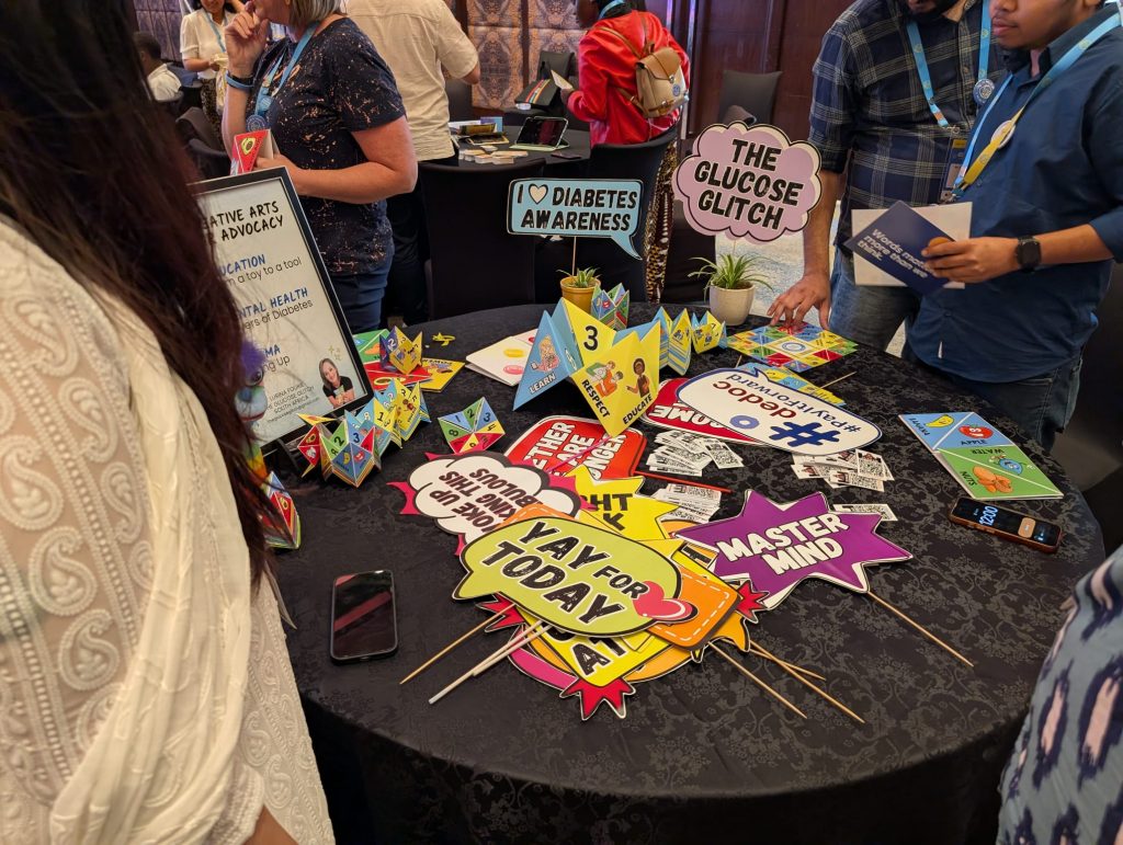 A group of conference attendees stands around a round table displaying a colourful diabetes awareness exhibit. The table is covered with interactive game pieces, educational materials, advocacy signs, and speech-bubble props with messages such as “The Glucose Glitch,” “Diabetes Awareness,” and “Yay for Today.” Participants wear conference lanyards and engage with the display, which highlights diabetes education, advocacy, and lived experiences through creative arts.