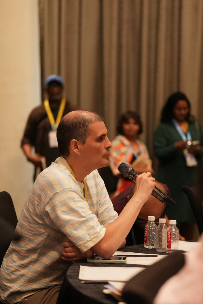 A medium shot focusing on a man with short grey hair speaking while standing at a conference table. He is wearing a grey Under Armour polo shirt, light-colored pants, a white smartwatch, and an event lanyard around his neck, and he is holding a white cane. In the blurred foreground, other attendees sit at the table, which holds water bottles and small cards. In the background, another man in a plaid shirt stands near the main stage area, which features lighting trusses and a large digital screen.