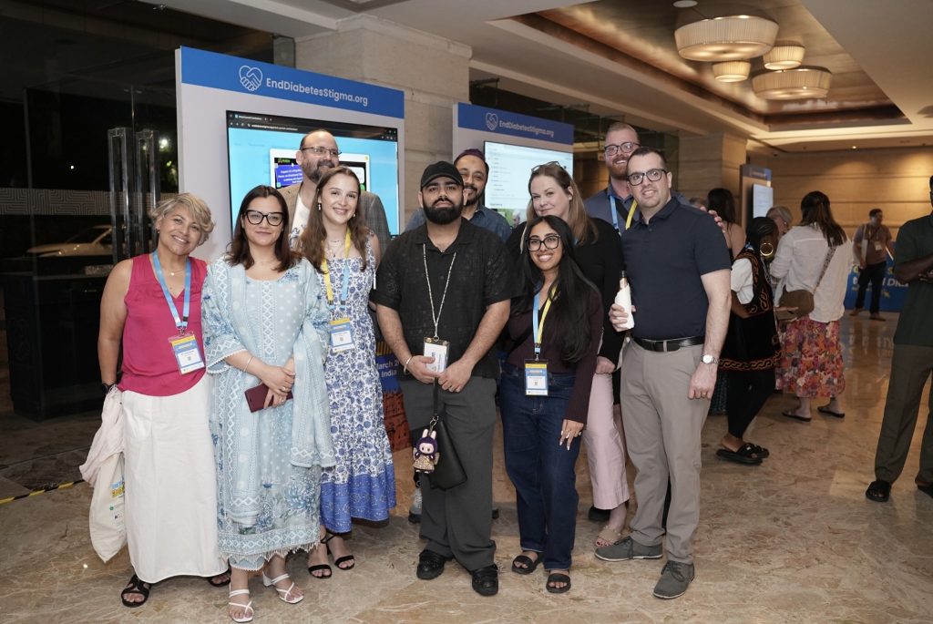 A group of nine people stand closely together smiling for a photo in a well-lit hotel or conference lobby with polished floors. They are dressed in casual and smart-casual attire, and several are wearing conference lanyards. In the background, modern digital displays feature the website "EndDiabetesStigma.org." Other event attendees can be seen mingling in the blurred background to the right.