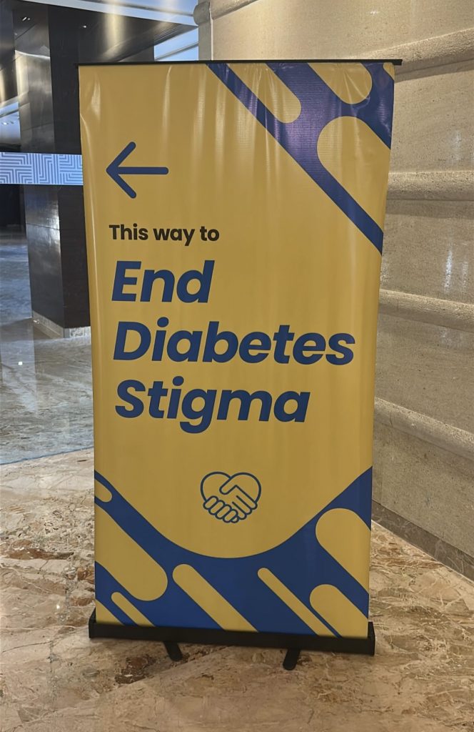 A bright yellow conference sign with bold blue text reading “This way to End Diabetes Stigma” and a blue arrow pointing left.
