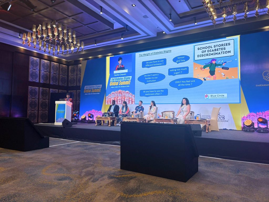 A wide shot of a brightly lit conference stage in a grand hall with decorative chandeliers. A presenter stands at a podium on the left beneath a screen showing her live video. Five panel members sit in chairs on stage to the right of the podium. The main backdrop screen displays the event title "End Diabetes Stigma Global Summit" alongside a large presentation slide detailing common stigmas, such as "eating too much sugar?" and "do you have to use the washroom often?", next to a graphic titled "School Stories of Diabetes Discrimination."