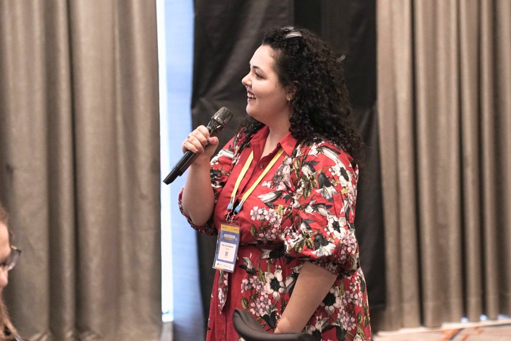 Linxi Mytkolli speaks into a microphone in front of conference attendees wearing a floral red dress, creating space for reflection and conversation around difficult topics in a calm and supportive setting.
