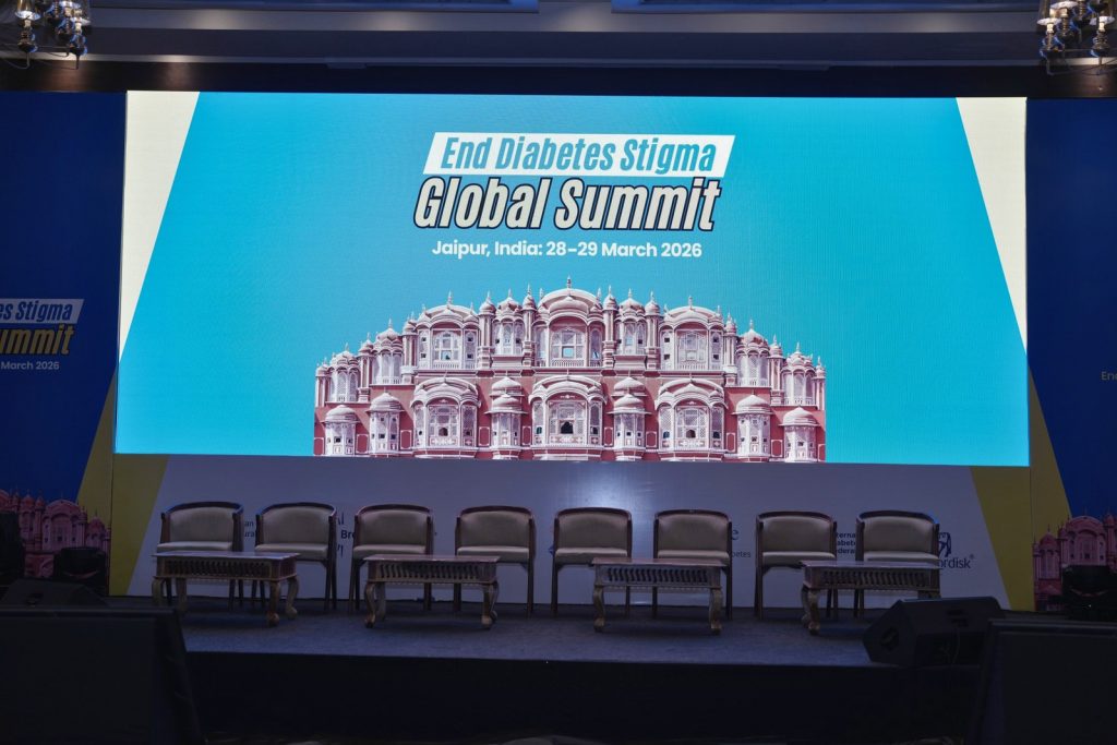 A conference stage set up for the "End Diabetes Stigma Global Summit" in Jaipur, India. A large digital screen displays the event title and an illustration of the Hawa Mahal palace against a bright blue background. Below the screen, eight empty chairs and four low wooden coffee tables are arranged on the stage for a panel discussion.
