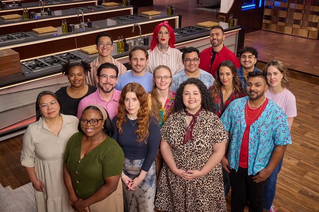 A high-angle group portrait of fifteen diverse people standing together inside a large, professional kitchen studio. They are arranged in multiple rows facing the camera with smiling or neutral expressions. The kitchen in the background features sleek wooden counters, stainless steel sinks, and cooktops. The individuals wear a variety of casual and semi-formal attire, including patterned shirts, dresses, glasses, and colorful hairstyles.